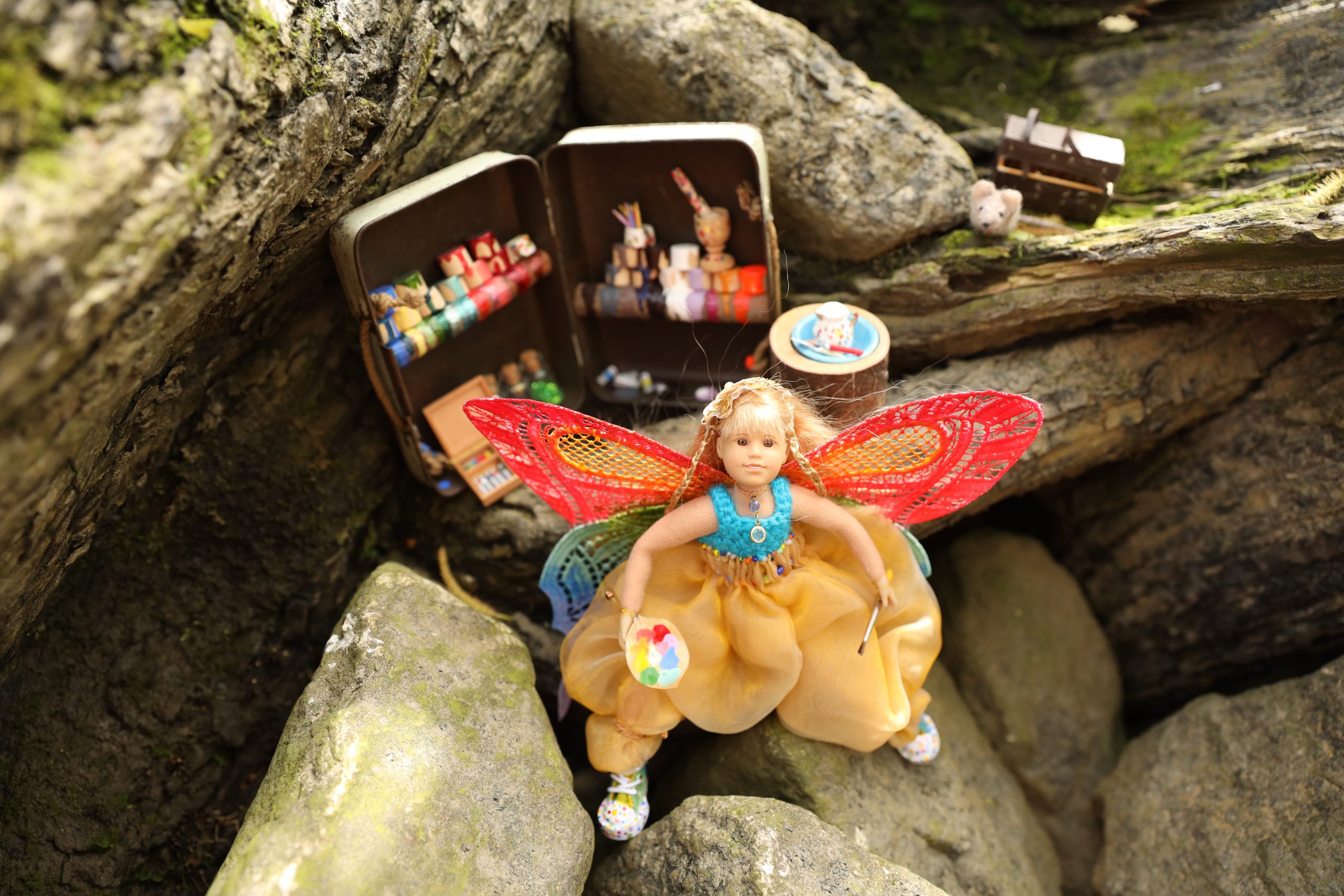eine bunt gekleidete Elfe steht draußen in der Natur vor ihrem Atelier. Farbtöpfe, Untensilien stehen hinter ihr. Sie hält einen Pinsel in der einen und eine Farbpalette auf einem Blatt in der anderen Hand.